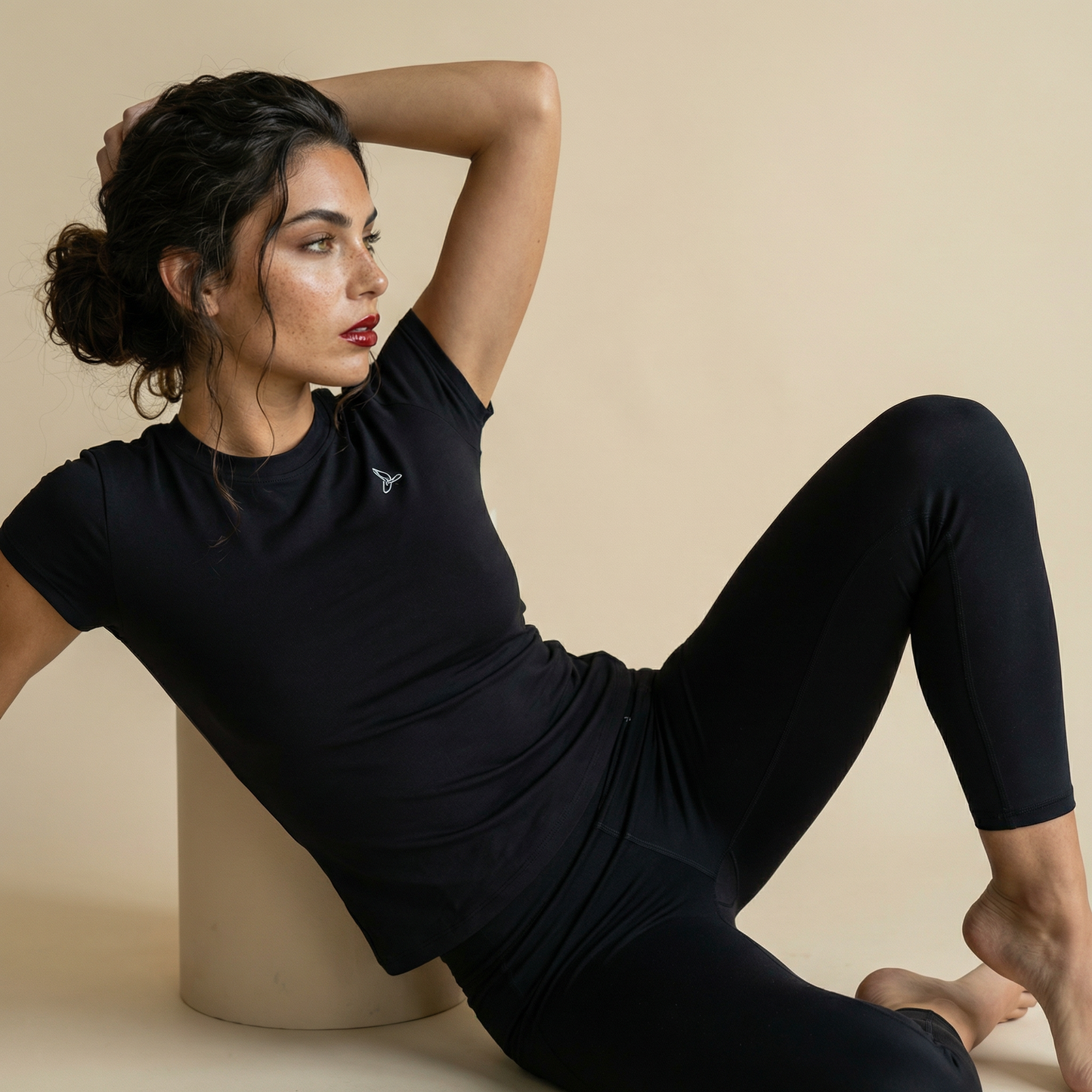 Woman seated wearing black short sleeve sculpting activewear top and black leggings, minimal luxury gym outfit styled in studio.